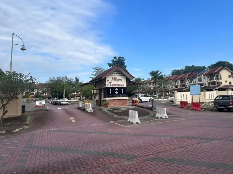 Exterior type: gated community; features: main entrance gate, brick facade at entrance, paved driveway, landscaped garden with trees, surrounding residential buildings, security barriers, street lamps, clear blue sky; style: modern.