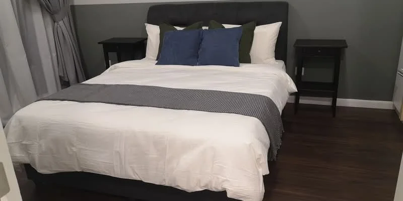 Bedroom features: king-size bed, upholstered headboard, white bedding, grey throw blanket, two bedside tables, blue decorative pillows, laminate flooring, curtains, wall with green accent, white walls; style: modern.