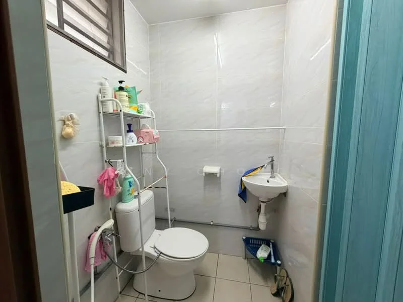 Bathroom features: white tiled walls, light grey tiled flooring, one-piece toilet, small pedestal sink, chrome faucet, shower caddy with toiletries, toilet paper holder, window for natural light, blue towel hanging, plastic storage basket.
