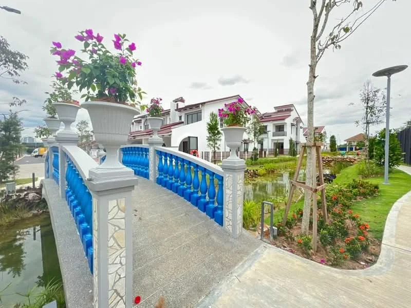 Exterior features: decorative blue bridge, flower pots with pink flowers, manicured landscaping, pathway with stone paving, water feature, trees with green foliage, residential buildings in background, street lighting, garden beds with flowers; style: modern.