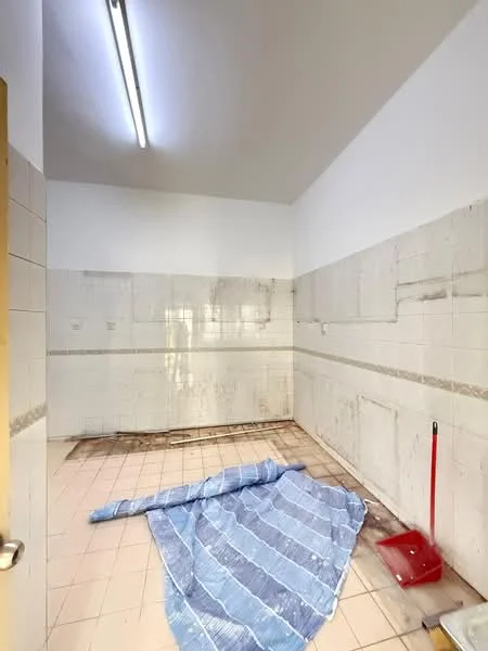 Room features: white tiled walls, beige tiled flooring, fluorescent ceiling lights, cleaning supplies visible, blue tarp on the floor, unfinished wall sections, no visible furniture, no windows.