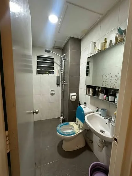 Bathroom features: white tiled walls, dark grey tiled flooring, walk-in shower with glass partition, chrome shower head, toilet with blue seat, single-sink vanity, mirror above sink, wall-mounted storage shelves, various toiletries on shelves, window for natural light, recessed ceiling lights; style: modern.