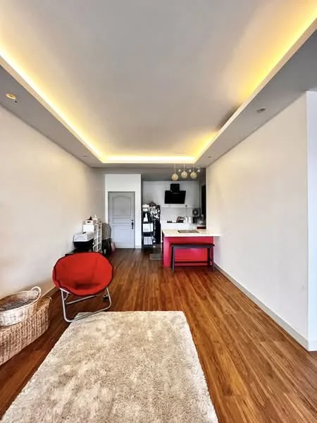 Living features: wood flooring, white painted walls, recessed downlights, ceiling with LED strip lighting, open layout, kitchen island, bar stools, TV mounted on wall, basket for storage, area rug, red accent chair; style: modern.