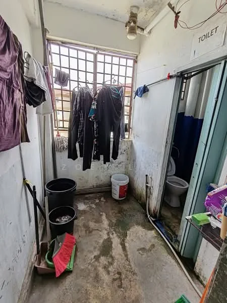 Laundry features: concrete flooring, white painted walls, clothes hanging on a line, bucket, plastic bin, toilet, partitioned area for toilet, window with metal bars, cleaning supplies, laundry basket.