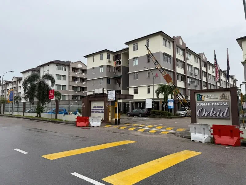 Exterior type: low-rise apartment block; features: grey painted facade, cream painted facade, multiple storeys, entrance gate with signage, landscaped area with palm trees, parking area visible, security guard house at entrance, road with yellow pedestrian crossing, surrounded by fencing, flag poles with flags, visible balconies, manicured lawn; style: modern.