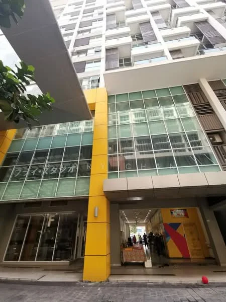 Exterior type: mid-rise condominium; features: glass curtain wall, yellow painted facade, recessed balconies, multiple floors visible, entrance with awning, large windows, commercial space at ground level, urban context with paved walkway; style: modern.