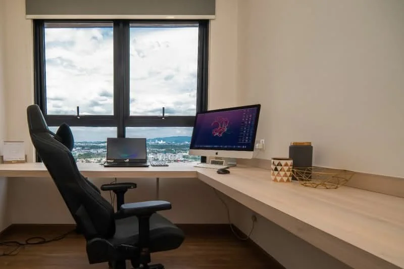 Study features: wooden desk, ergonomic office chair, desktop computer, laptop, window with city view, light-colored wall, laminate flooring, decorative storage basket; style: modern; view: city.