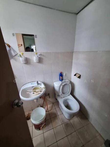 Bathroom features: white ceramic toilet, white bidet, wall-mounted sink, beige tiled walls, light beige floor tiles, toilet paper holder, cleaning supplies visible, bucket on the floor.