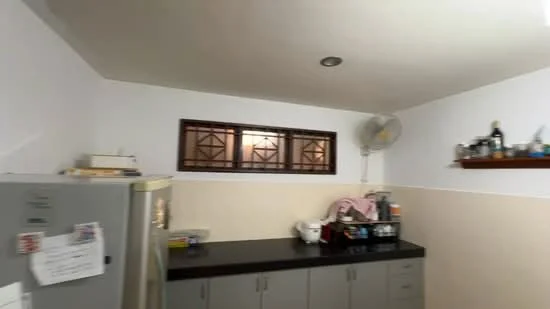 Kitchen features: grey cabinetry, black countertop, stainless-steel fridge, wall-mounted ceiling fan, shelf with various items, window with decorative grille, light-colored walls, tile backsplash.