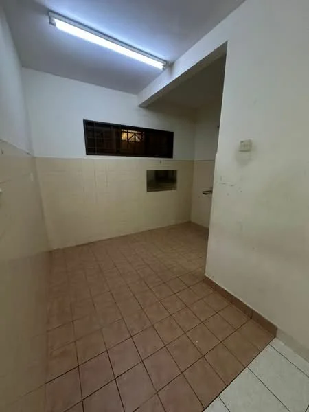 Room features: white painted walls, brown tiled flooring, window with grille, sink area, fluorescent ceiling light, corner wall, plain ceiling, tile cladding on lower walls.