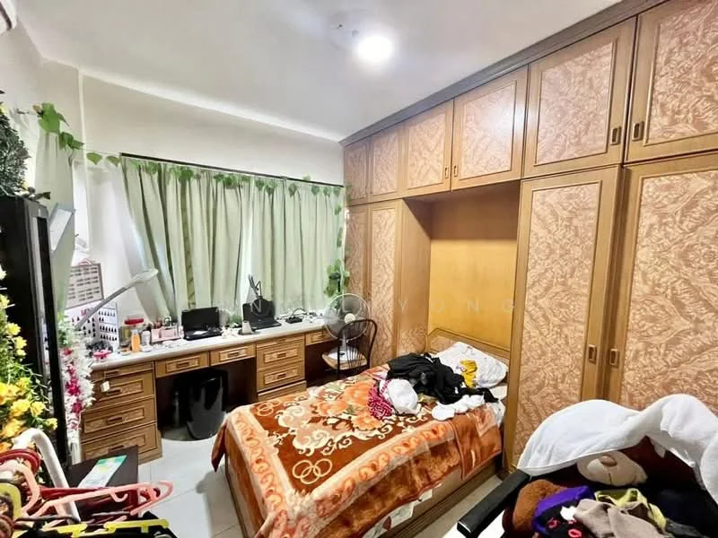 Bedroom features: wooden flooring, built-in wardrobe with wood finish, queen-size bed with patterned bedspread, study desk with drawers, desk lamp, chair, ceiling light, green curtains, wall-mounted fan, decorative plants, clothes on bed.
