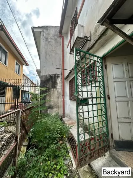 Exterior features: green metal gate, concrete pathway, exposed brick wall, air conditioning unit mounted on wall, overgrown vegetation, adjacent building visible, fenced area.