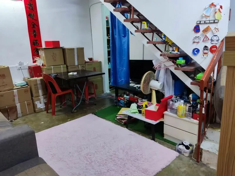 Living features: grey sofa, pink area rug, wooden dining table, red plastic chairs, green carpet, exposed concrete flooring, staircase with wooden railing, storage boxes, fan, wall-mounted decorations, shelves with various items.