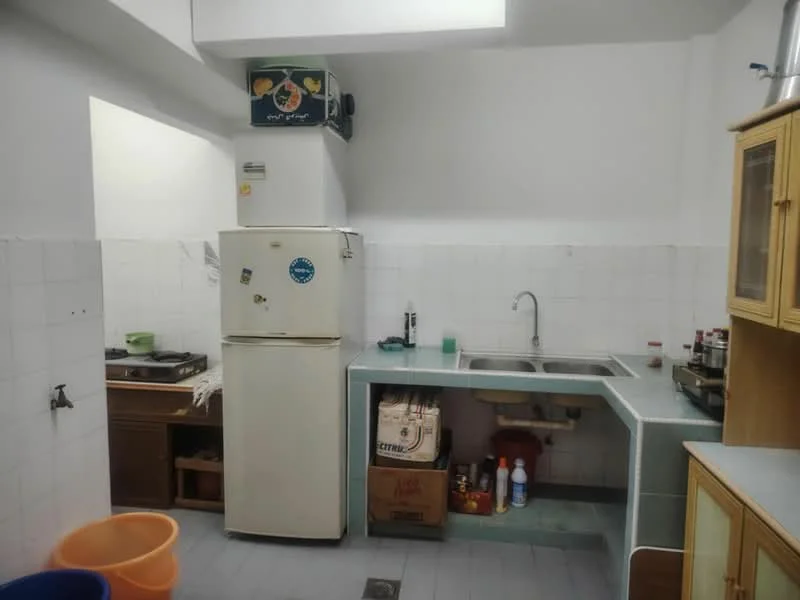 Kitchen features: white tiled walls, light grey countertop, single-bowl stainless-steel sink, chrome faucet, white fridge, freestanding gas stove, wood cabinetry, orange plastic trash bin, storage boxes under sink, grey tiled flooring.