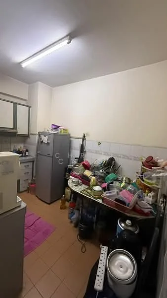 Kitchen features: grey cabinetry, stainless-steel fridge, white tiled backsplash, ceramic tile flooring, overhead fluorescent lighting, countertop with cluttered utensils, sink with faucet, multiple plastic containers, kitchen table with various items, pink mat on floor.