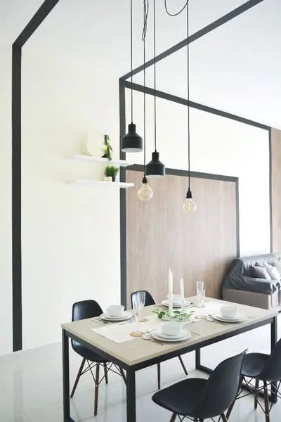 Dining features: dining table with black metal frame, 4 black dining chairs with wooden legs, white plates on the table, glassware on the table, candles on the table, pendant lights with black shades, white walls with black trim, wood panel accent wall, polished tile flooring, shelf with decorative items; style: modern.