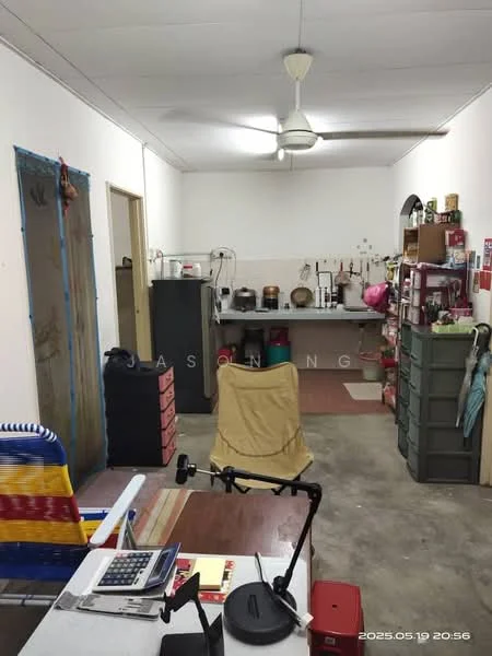 Room features: white painted walls, concrete flooring, ceiling fan, kitchen area with cabinets, stainless-steel sink, gas stove, wooden countertop, refrigerator, storage boxes, dining table, office desk, office chair, various kitchen utensils, wall-mounted shelves.