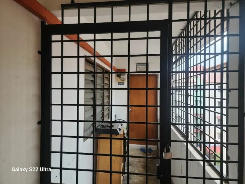 Room features: metal security gate, grid pattern on gate, wooden door, concrete flooring, white walls, window with metal grilles, exposed electrical wiring, light fixture on ceiling.