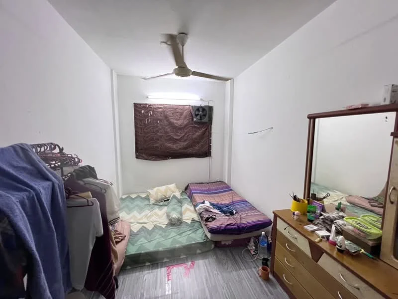 Bedroom features: two single beds with bedding, wooden dresser with mirror, ceiling fan, wall-mounted air conditioning unit, white painted walls, grey tiled flooring, brown window curtain, clothes rack with hanging clothes, decorative items on dresser, bottled water on floor.