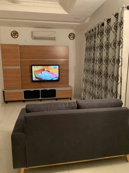 Living features: 3-seater fabric sofa in dark grey, TV console with wooden finish, wall-mounted TV, recessed ceiling lights, white walls, floor-to-ceiling curtains with geometric pattern, air conditioning unit, tile flooring, decorative wall accents; style: modern.