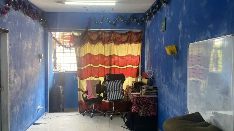 Room features: blue painted walls, white ceiling, tile flooring, large window with metal grille, red and yellow patterned curtains, two office chairs, black bookshelf, whiteboard, decorative garland, stack of books, small table, electrical outlet, wall-mounted light fixture.