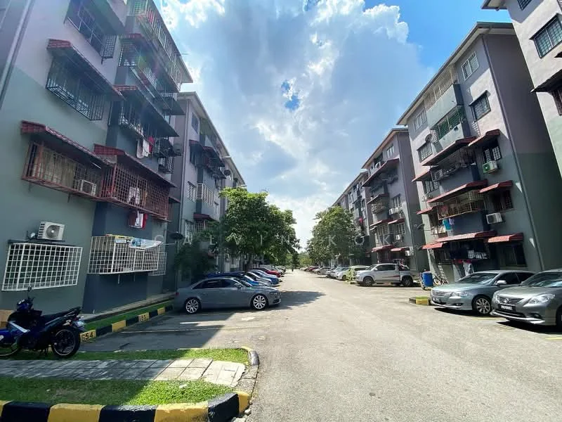 Exterior type: low-rise apartment block; features: grey painted facade, balconies with metal railings, air conditioning units visible, parking spaces visible, landscaped green area, paved driveway, surrounded by trees, two-storey building, multiple vehicle parking, cloudy sky; style: modern.