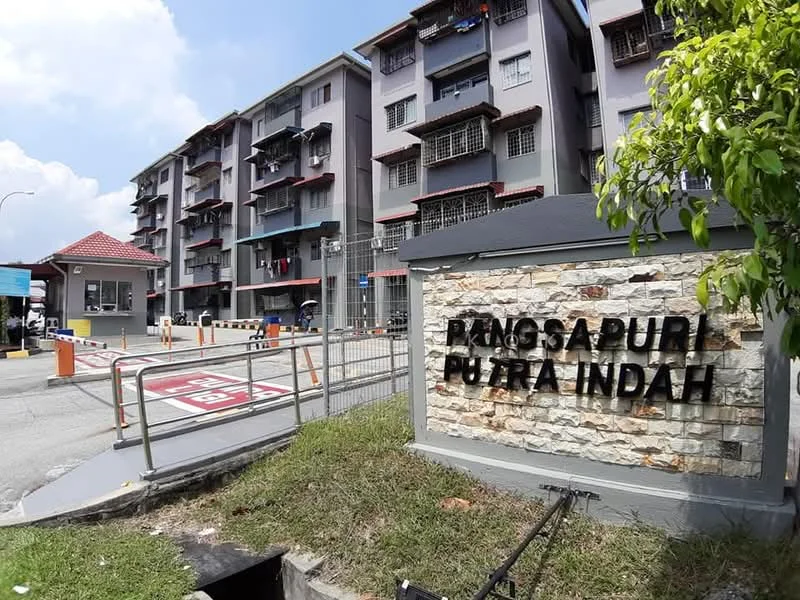 Exterior type: low-rise apartment block; features: grey painted facade, stone cladding at entrance sign, security guard house at entrance, gated entry, multiple balconies visible, manicured lawn, paved walkway, parking area visible; style: modern.