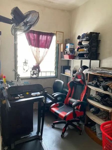 Study features: desk with computer setup, black office chair, red and black gaming chair, wall-mounted fan, window with sheer curtains, shelves with storage boxes, clothes on open shelving, grey flooring, decorative certificate on wall, red laundry basket.