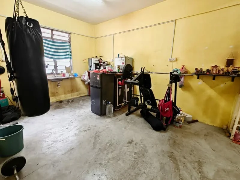 Room features: yellow painted walls, concrete flooring, window with striped curtains, refrigerator, weight bench, dumbbells, punching bag, various containers and items on shelves, plastic bucket, decorative items on shelves.