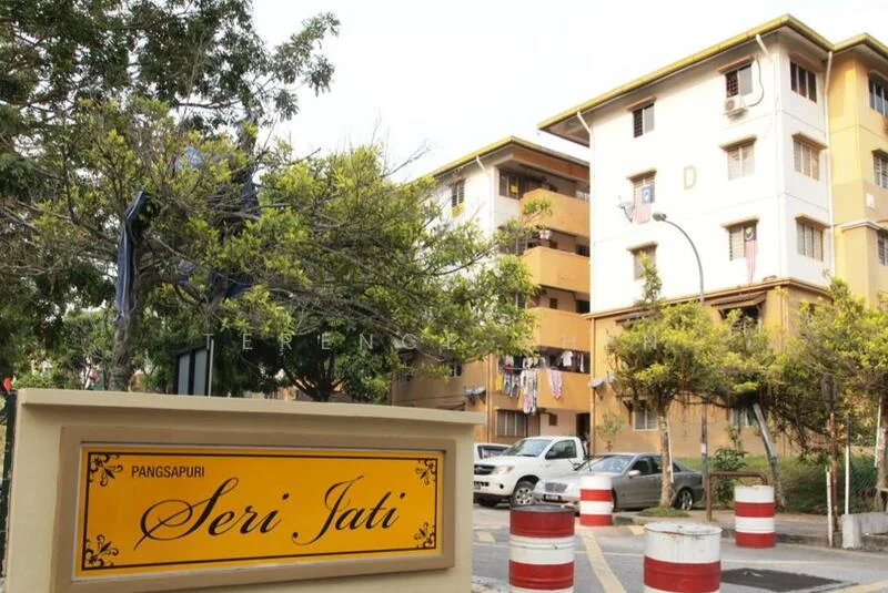 Exterior type: low-rise apartment block; features: yellow painted facade, 3-storey building, balconies with railings, visible laundry on balconies, landscaped greenery, parking area, road with traffic barriers; style: modern.