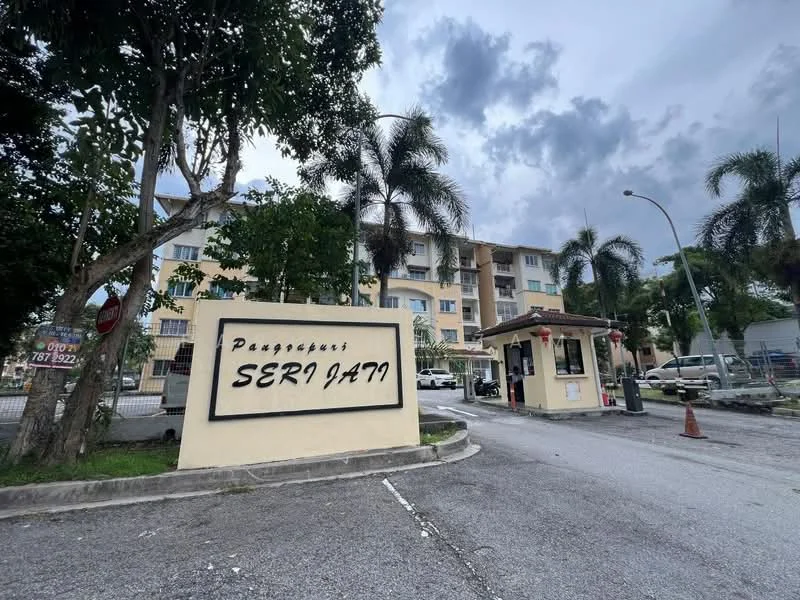 Exterior type: low-rise apartment block; features: main entrance gate, guard house at entrance, palm trees, manicured landscaping, parking area visible, multiple storeys, signage with property name, surrounded by greenery; style: modern.