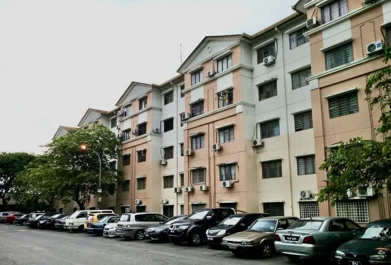 Exterior type: low-rise apartment block; features: cream painted facade, light grey painted facade, multiple air conditioning units visible, parking lot with various vehicles, surrounded by trees, street lighting, multiple storeys, balconies visible; style: modern.