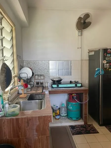 Kitchen features: single-bowl stainless-steel sink, chrome faucet, gas stove with blue burner, brown wooden countertop, white tiled backsplash, grey tiled flooring, refrigerator with stainless-steel finish, wall-mounted fan, green gas cylinder, cleaning supplies on countertop, decorative mat on floor.