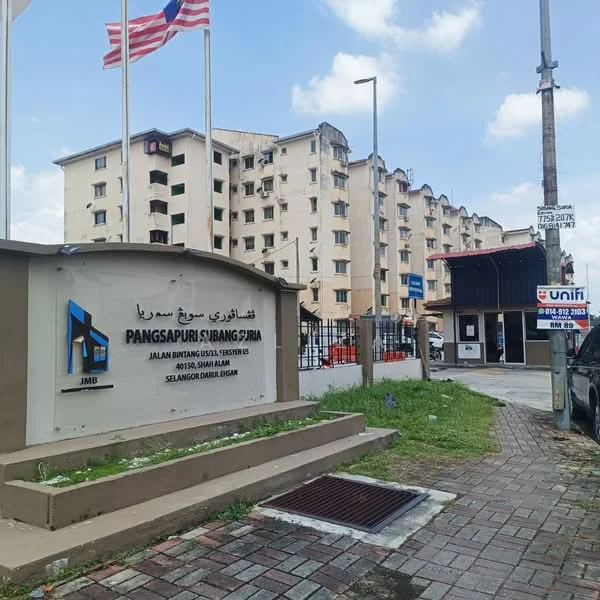 Exterior type: low-rise apartment block; features: cream painted facade, multiple storeys, balconies on upper floors, security guardhouse at entrance, paved walkway, surrounded by mature greenery, flag poles with Malaysian flags, visible signage for the building, parking area, street view with utility poles; style: modern.