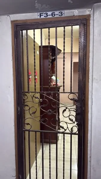 Room features: metal security door with decorative design, light beige walls, tiled flooring, wooden furniture visible in background, wall-mounted decorations, doorway leading to interior space.