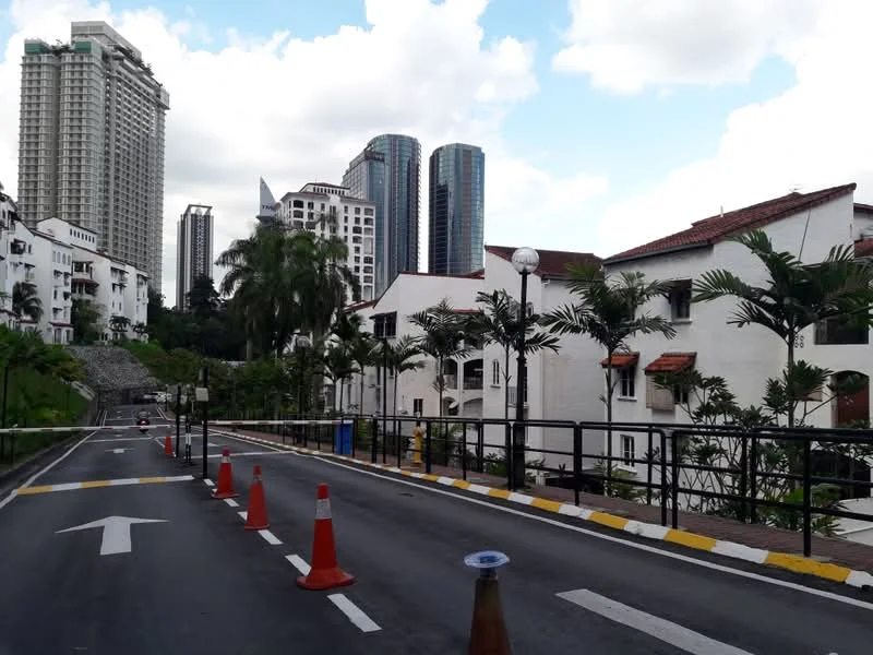 Exterior type: linked terrace house; features: white painted facade, red tiled roof, palm trees, manicured landscaping, guard house at entrance, surrounded by mature greenery, multi-storey high-rise buildings in background, paved road, traffic cones, street lamps; style: modern.