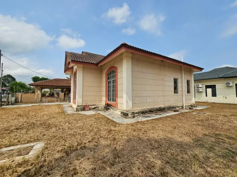 Exterior type: detached bungalow; features: cream painted facade, red trim around windows and doors, pitched tile roof, single-storey, surrounded by dry grass, visible air conditioning units, concrete pathway, mature trees in background; style: traditional.