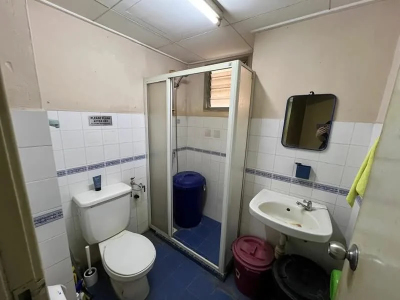 Bathroom features: white tiled walls, blue tiled flooring, shower cubicle with glass door, toilet, single-sink vanity with mirror, blue trash bin, pink trash bin, window for natural light, towel rack, soap dispenser.