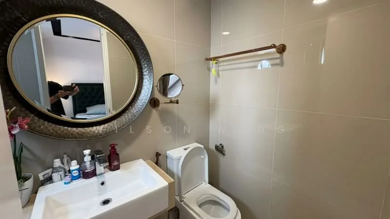 Bathroom features: white ceramic toilet, single-sink vanity with white cabinet, round mirror with decorative frame, wall-mounted towel bar in bronze, grey tiled walls, recessed lighting, chrome faucet, bottle of hand soap, bottle of lotion, decorative plant, toilet paper holder; style: modern.