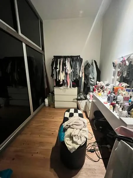 Bedroom features: wooden flooring, large mirrored wardrobe, clothing rack with various garments, white dresser, vanity table with cosmetics, black and white striped ottoman, overhead lighting, wall with neutral paint.