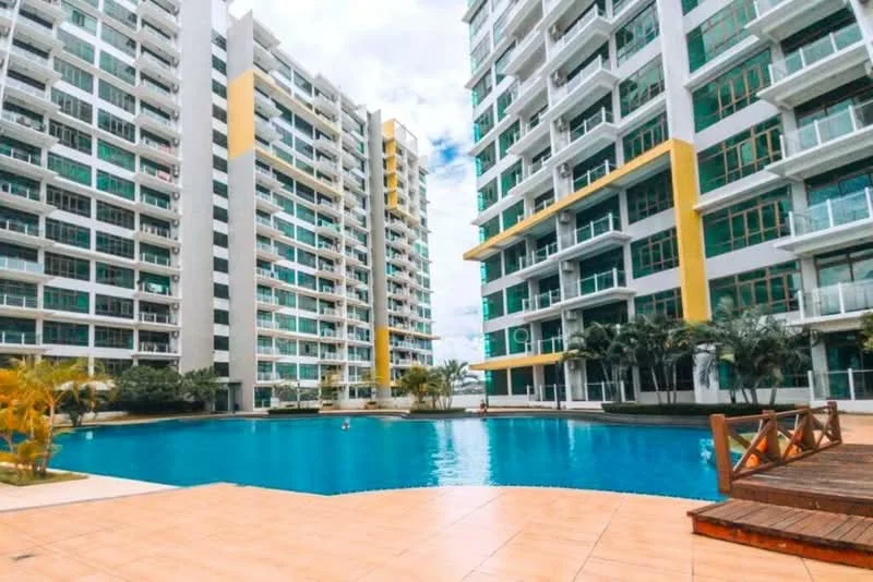 Exterior type: high-rise condominium; features: swimming pool, pool deck with loungers, surrounded by palm trees, manicured landscaping, modern glass facade, yellow accent panels, multiple balconies, outdoor wooden deck, water feature, open-air common area; style: modern.