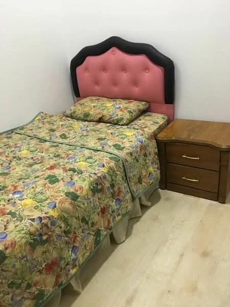 Bedroom features: queen-size bed with floral bedding, pink upholstered headboard, wooden bedside table, two-drawer bedside table, laminate flooring, white painted walls, decorative pillows; style: traditional.