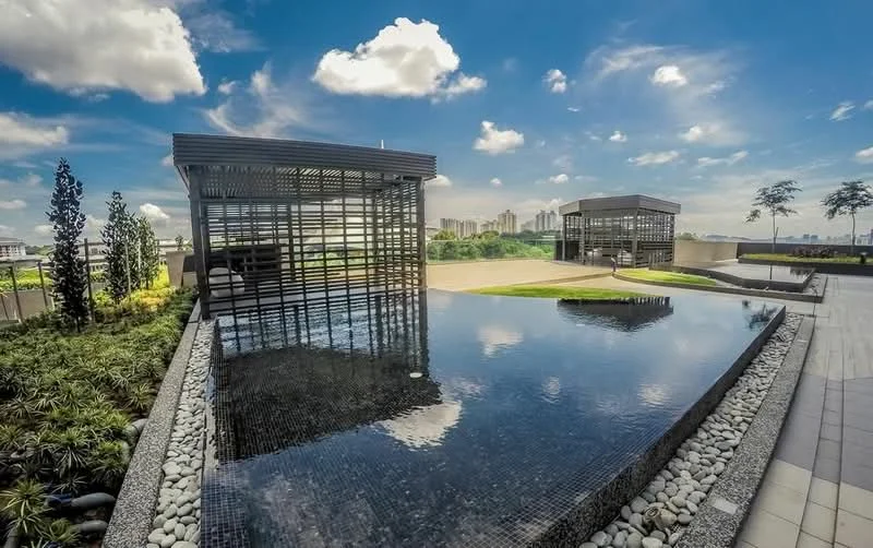 Exterior type: landscape garden; features: infinity pool, pool deck with loungers, manicured lawn, modern pavilion structures, decorative stone landscaping, surrounding greenery, urban skyline in background, clear blue sky; style: modern.
