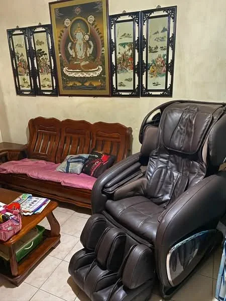 Living features: brown leather massage chair, wooden sofa with backrest, pink and blue cushions, decorative wall art with religious theme, framed artwork with traditional motifs, ceramic floor tiles, wooden coffee table, magazine on coffee table; style: traditional.