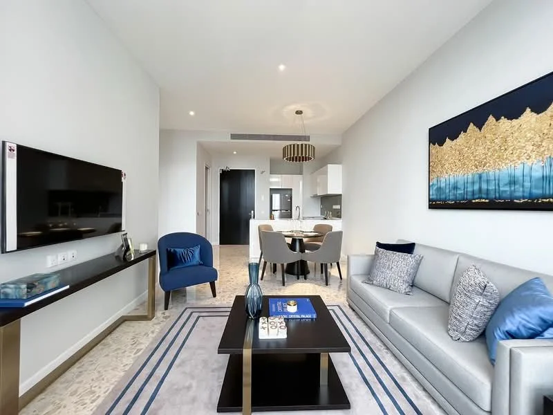 Living features: grey fabric sofa, blue accent chairs, coffee table with black surface, TV console with wall-mounted TV, decorative wall art with gold and blue tones, recessed ceiling lights, neutral wall colour, area rug with geometric pattern, dining area with round table and chairs in background; style: modern.