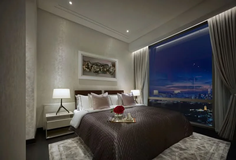 Bedroom features: king-size bed with brown quilted cover, upholstered headboard, two bedside lamps with white shades, large floor-to-ceiling window with city view, curtains in light grey, decorative wall art above the bed, dark wood bedside tables, area rug with geometric pattern, recessed ceiling lights, cream painted walls, dark wood flooring; style: modern; view: city.