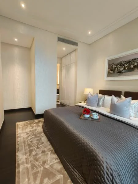 Bedroom features: king-size bed with grey quilted cover, upholstered headboard, bedside lamps, decorative pillows, area rug with geometric pattern, built-in wardrobe with sliding doors, wall-mounted artwork, recessed ceiling lights, dark wood flooring, neutral painted walls; style: modern. Room features: narrow hallway leading to another room, light-colored walls, recessed lighting.