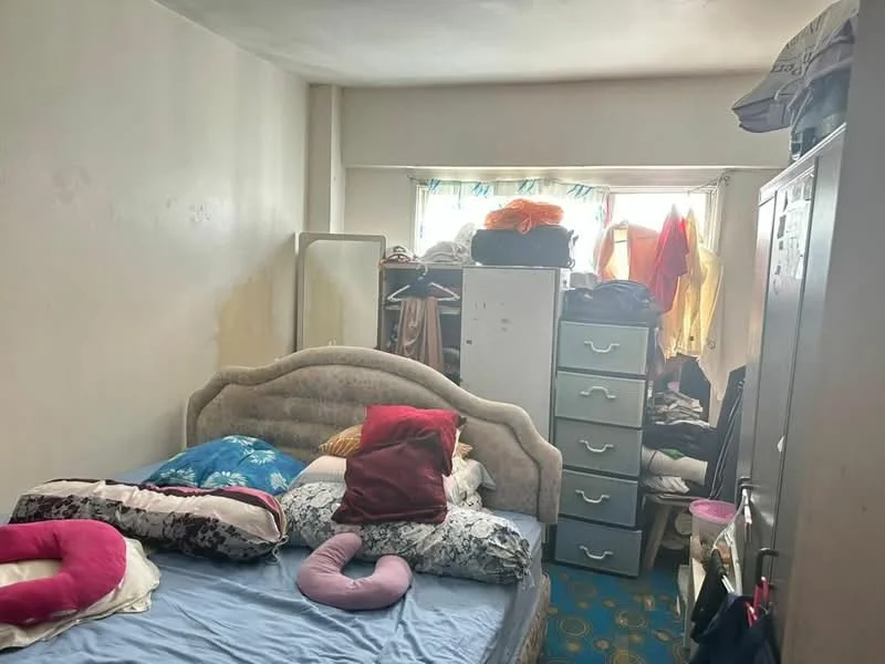 Bedroom features: double bed with light blue bedding, upholstered headboard, decorative pillows in various colors, large window with sheer curtains, wardrobe with sliding doors, chest of drawers, clothes piled on top of wardrobe, carpeted flooring with blue pattern, pink neck pillow, cluttered storage area, mirror leaning against the wall.