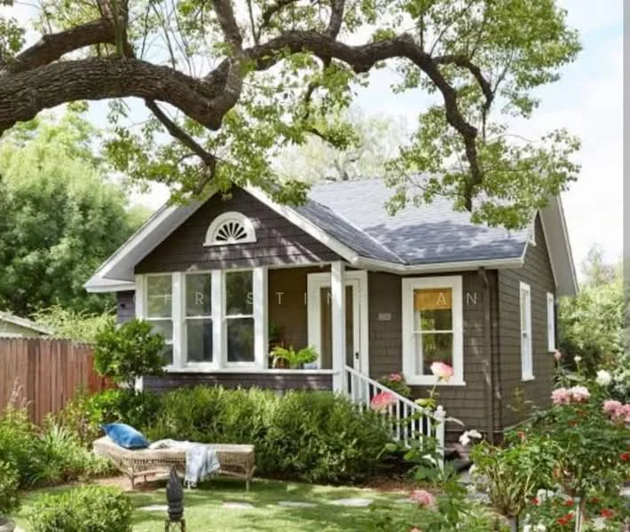 Exterior type: detached bungalow; features: wooden facade with dark brown paint, pitched roof with shingles, front porch with white railing, large front windows, decorative gable vent, manicured lawn, flower beds with roses, mature trees providing shade, stone pathway leading to entrance, outdoor seating area with cushions; style: traditional.