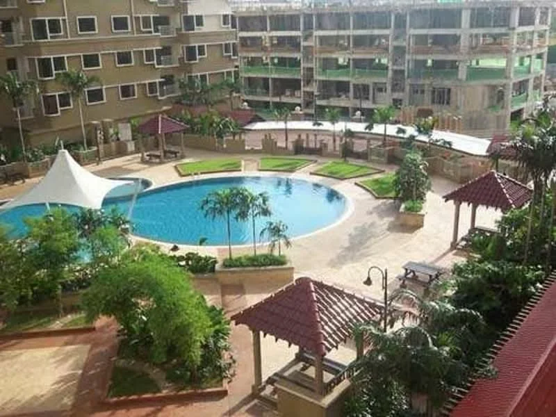 Exterior type: mid-rise condominium; features: swimming pool with circular shape, pool deck with loungers, landscaped garden with manicured lawns, pavilions with tiled roofs, tropical landscaping with palm trees, walking paths around the pool, outdoor seating area, covered gazebo, surrounding greenery; style: modern.